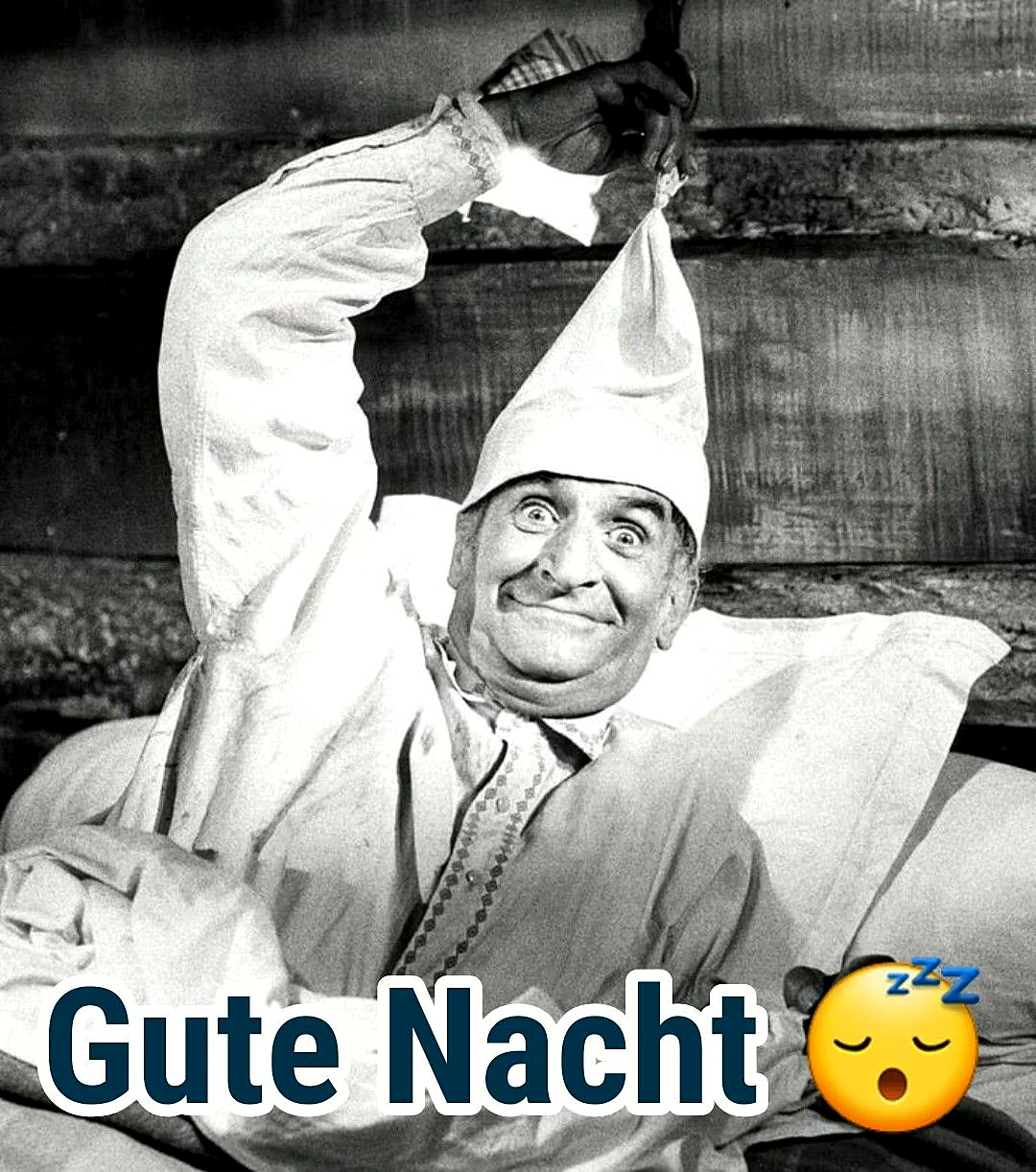 Schwarz-weißes Bild eines Mannes mit breitem, komischem Grinsen und spitzer Schlafmütze. Er zieht die Mütze hoch, liegt im Bett. Der Text 'Gute Nacht' mit schlafendem Emoji vermittelt eine humorvolle Abendstimmung.