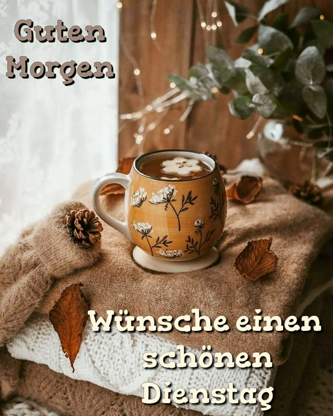 Eine gemütliche Szene zeigt eine verzierte Teetasse auf weichen Strickpullovern. Herbstblätter, Kiefernzapfen und funkelnde Lichterketten im Hintergrund schaffen eine warme, ruhige Morgenstimmung. Ein Eukalyptuszweig ergänzt das Motiv.