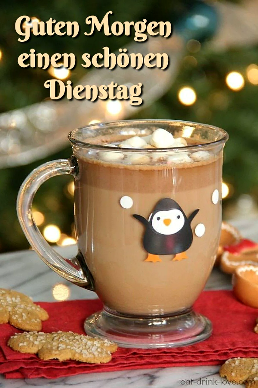 Ein gemütlicher gläserner Becher gefüllt mit Kakao und Mini-Marshmallows, geschmückt mit einem Pinguin, steht auf einem roten Tuch neben zuckerbestreuten Keksen vor einem weihnachtlich leuchtenden Baum.