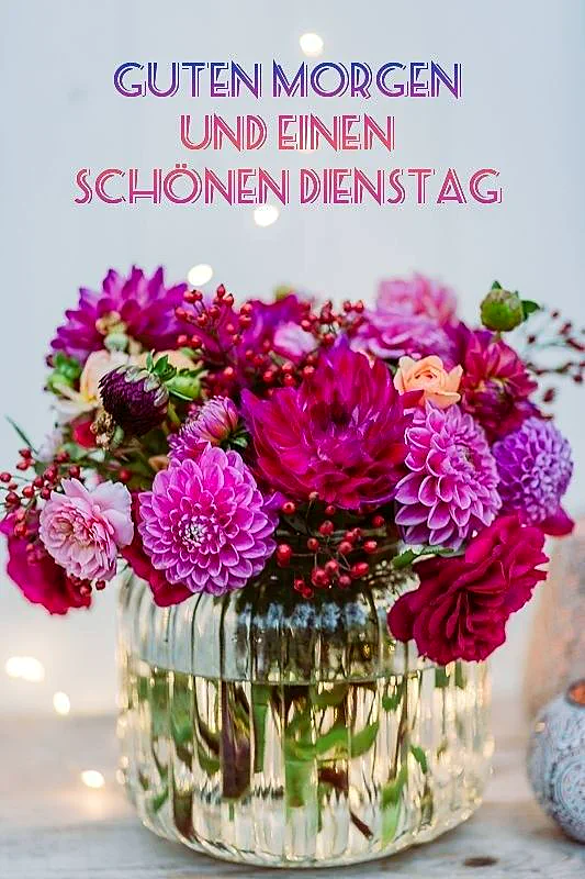 Ein prachtvoller Blumenstrauß mit lila und pinkfarbenen Dahlien sowie roten Beeren in einer gerippten Glasvase. Der helle, weichgezeichnete Hintergrund mit warmen Bokeh-Lichtern schafft eine freundliche, motivierende Morgenstimmung.